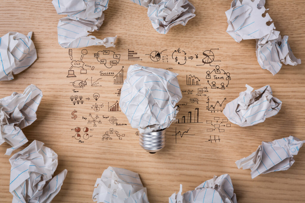 A table covered in equations is littered with crumpled up pieces of paper, one paper is in the shape of a lightbulb.